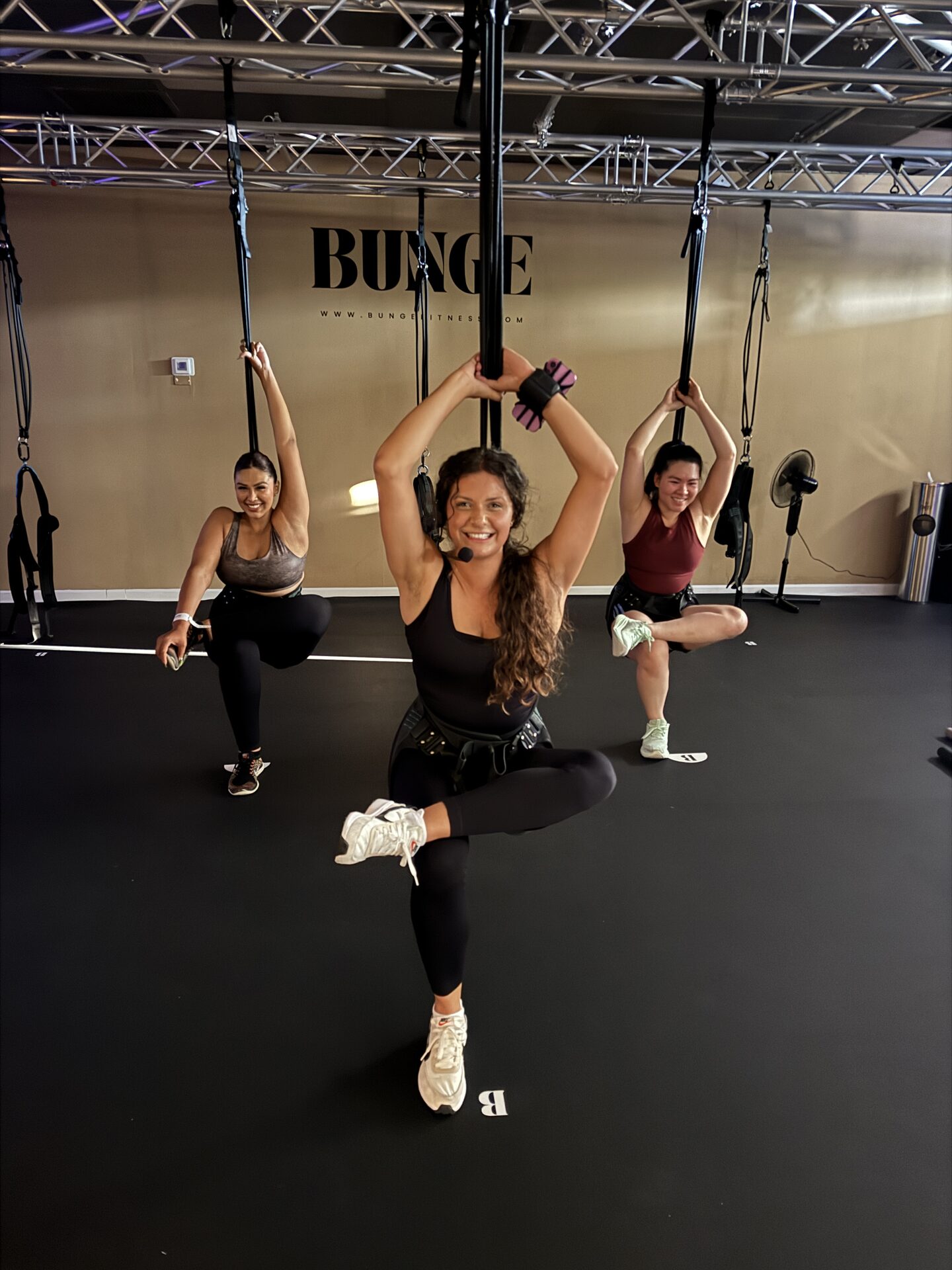 Instructor with a headset mic leading a bungee fitness class in front of the BUNGE logo wall in Huntington Beach