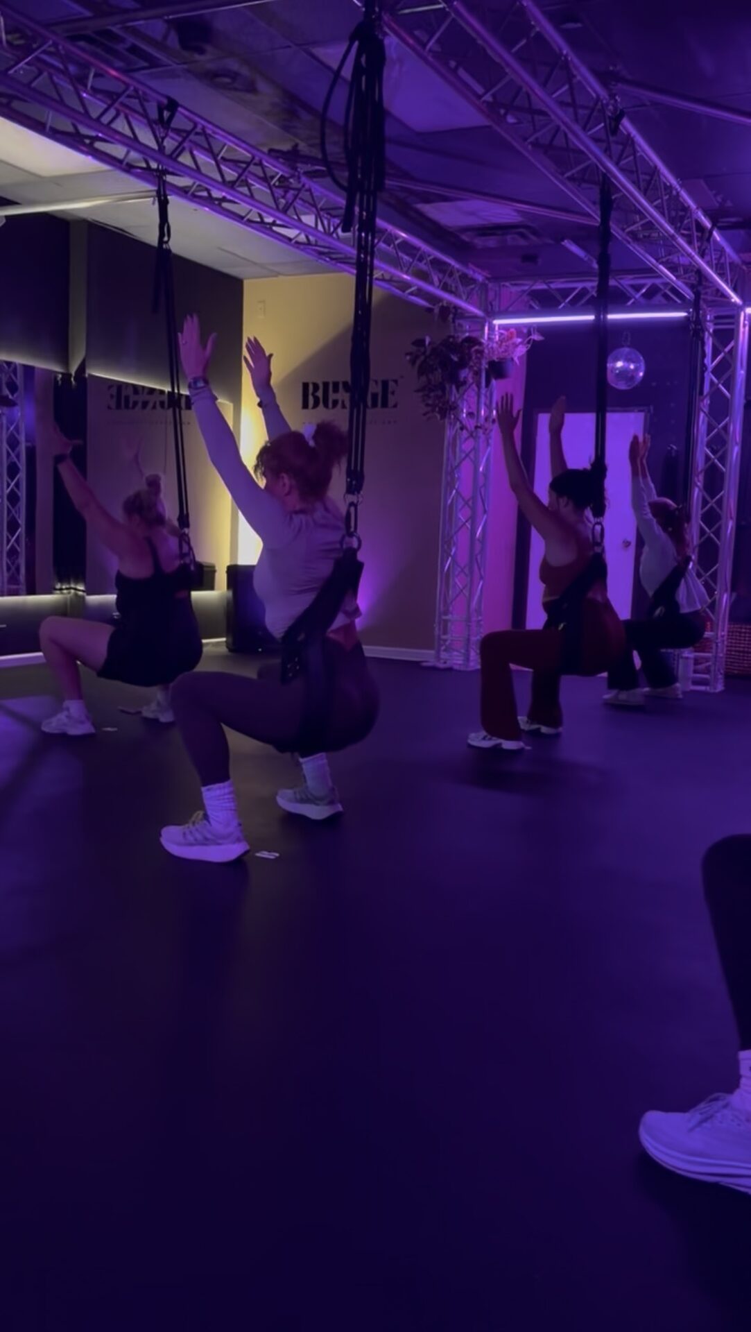Friends seated in bungee swing harnesses under purple light and a disco ball during a private party at BUNGE Fitness