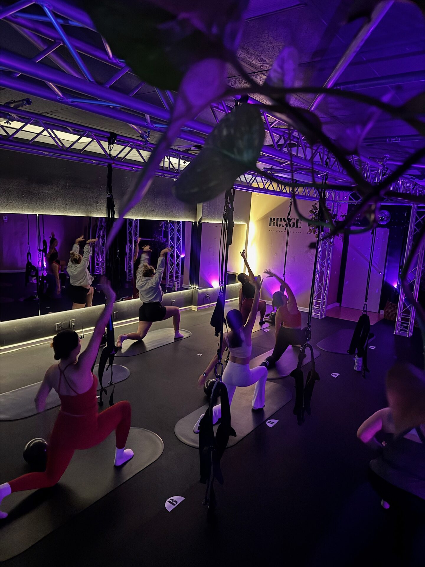 Students holding a deep lunge reach under purple lighting during a bungee fitness class at BUNGE Fitness in Huntington Beach
