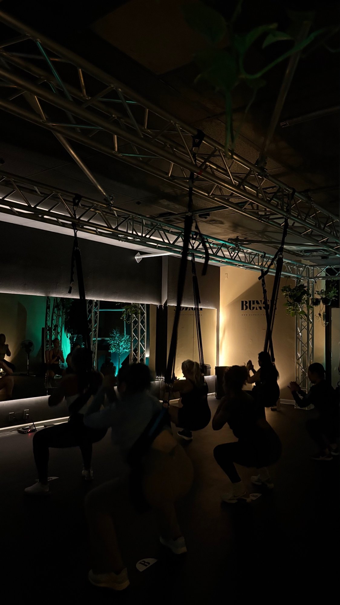 A group squat sequence under teal lighting during a bungee fitness class at BUNGE Fitness