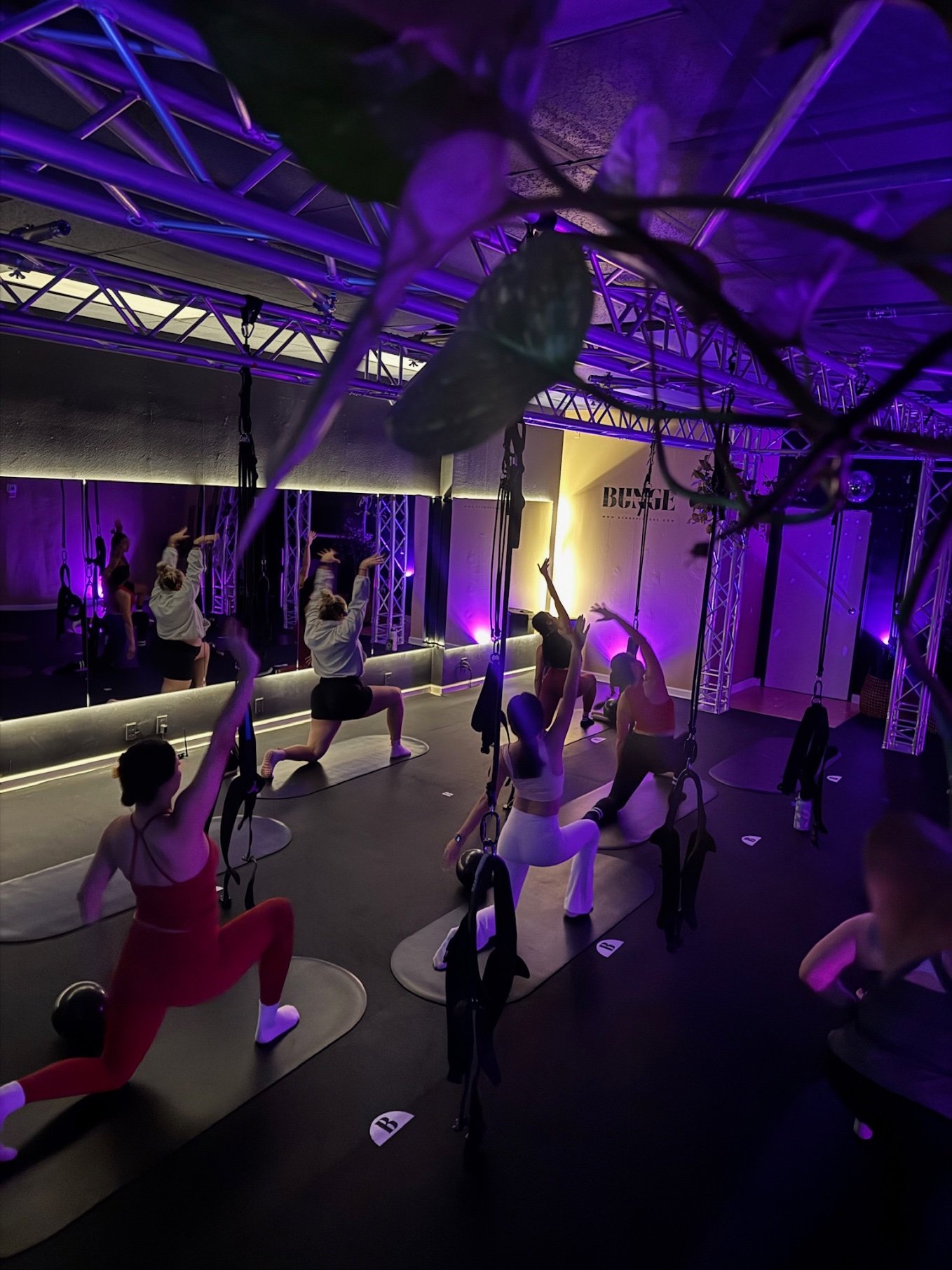 Students holding a deep lunge reach under purple lighting during a bungee fitness class at BUNGE Fitness in Huntington Beach